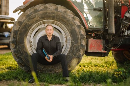 Valmiera, Letonia - 17 de agosto de 2024 - Un hombre está en cuclillas junto a un gran neumático tractor, sosteniendo un portapapeles, con maquinaria agrícola en el fondo.