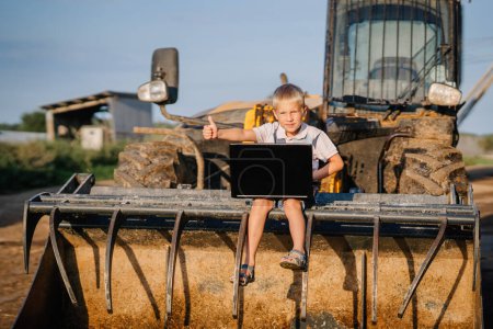 Valmiera, Letonia - 17 de agosto de 2024 - Un niño pequeño se sienta en un cubo de tractor, dando un pulgar hacia arriba y mirando la pantalla de un portátil.