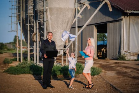 Valmiera, Lettland - 17. August 2024 - Ein Mann mit Laptop, eine Frau mit Klemmbrett und ein Kind werfen Papierflugzeuge auf einen Bauernhof mit Silos.