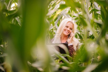 Valmiera, Lettland - 17. August 2024 - Eine lächelnde Frau mit blonden Haaren führt ein Telefongespräch, während sie einen Laptop in einem Maisfeld hält..