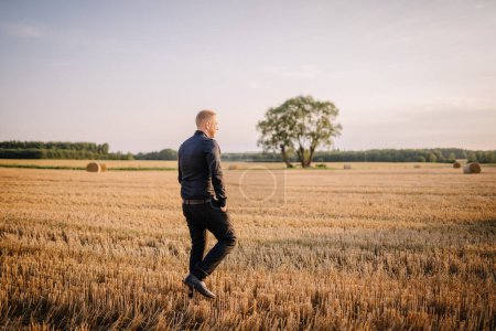 Valmiera, Lettonie - 17 août 2024 - Un homme en tenue d'affaires traverse un champ récolté avec des balles de foin et un arbre solitaire en arrière-plan.