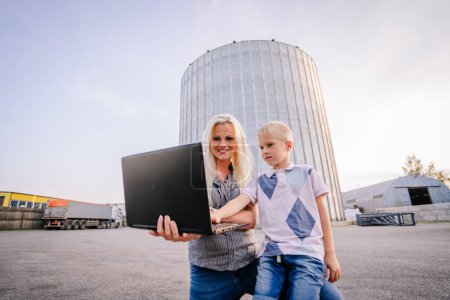 Foto de Valmiera, Letonia - 17 de agosto de 2024 - Una mujer sonriente muestra una pantalla de computadora portátil a un joven afuera cerca de grandes silos industriales. - Imagen libre de derechos