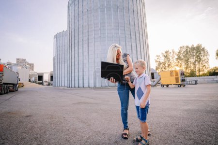 Téléchargez les photos : Valmiera, Lettonie - 17 août 2024 - Une femme souriante tenant un ordinateur portable montre quelque chose à un jeune garçon à ses côtés, tous deux debout dans un cadre industriel avec de grands silos et un coucher de soleil doré derrière eux. - en image libre de droit