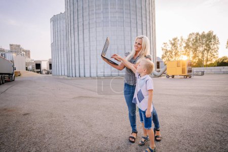 Valmiera, Latvia - August 17, 2024 - A woman and boy, likely mother and son, look at a laptop screen together in an industrial area with silos in the background at sunset.