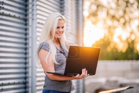 Valmiera, Lettland - 17. August 2024 - Eine lächelnde Frau mit langen blonden Haaren benutzt draußen einen Laptop mit Metallstrukturen im Hintergrund und warmem Sonnenlicht, das von der Seite flackert..