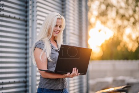 Valmiera, Lettland - 17. August 2024 - Eine lächelnde Frau mit langen blonden Haaren benutzt draußen einen Laptop mit Metallstrukturen im Hintergrund und warmem Sonnenlicht, das von der Seite flackert..