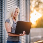 Valmiera, Latvia - August 17, 2024 - A smiling woman with long blonde hair is using a laptop outside with metal structures in the background and warm sunlight flaring from the side.