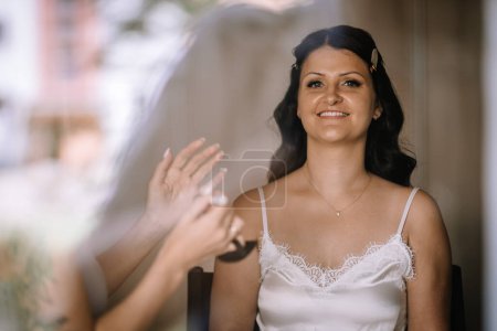 Valmiera, Lettland - 28. Juli 2024 - Eine Braut sitzt lächelnd da, während eine Visagistin ihr Make-up mit Spray einsprüht.