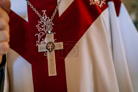Valmiera, Lettonie- 28 juillet 2024 - Gros plan d'un pendentif en croix d'argent sur une chaîne au-dessus de vêtements religieux avec un dessin rouge et blanc, indicatif d'un clergé ou d'un prêtre.