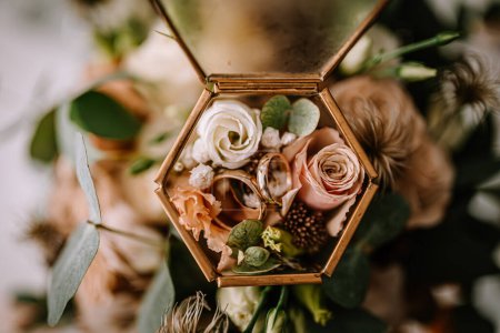 Valmiera, Letonia 28 de julio de 2024 - Los anillos de boda descansan en una caja de vidrio hexagonal rodeada por un arreglo de flores, destacando un momento de simbolismo romántico y compromiso..