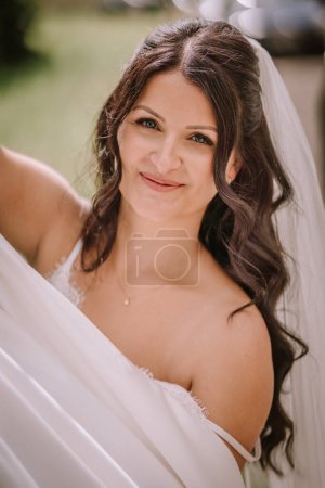 Photo for Valmiera, Latvia- July 28, 2024 - A smiling bride with a veil and dark wavy hair is holding her white wedding dress, looking joyous on her wedding day with a soft-focus background. - Royalty Free Image