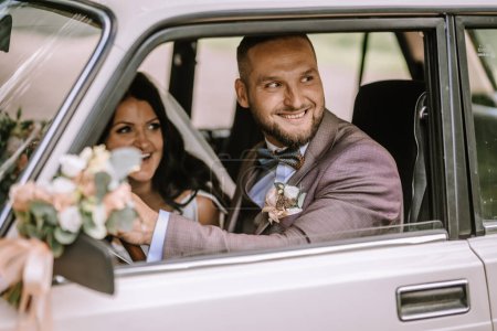 Foto de Valmiera, Letonia 28 de julio de 2024 - Novia y novio sonriendo dentro de un coche clásico, novio al volante, novia con un ramo, ambos mirándose alegremente el uno al otro. - Imagen libre de derechos