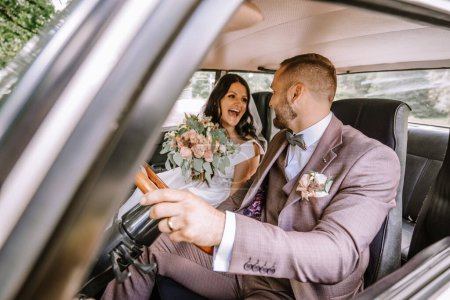Valmiera, Latvia- July 28, 2024 - Laughing bride and groom sit in a vintage car, the bride holding a bouquet, as the groom playfully pretends to drive.
