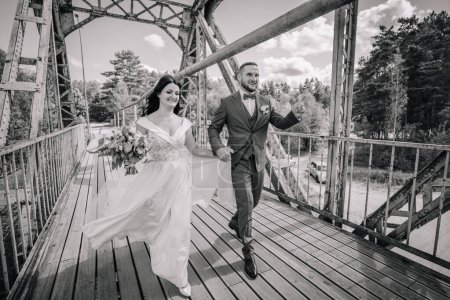 Téléchargez les photos : Valmiera, Lettonie- 28 juillet 2024 - Mariée et marié se tenant la main et courant sur un pont de fer, riant joyeusement sur une photo en noir et blanc. - en image libre de droit
