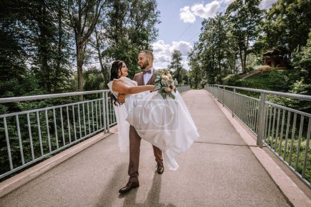 Photo for Valmiera, Latvia- July 28, 2024 - A groom carries his bride across a bridge, while a couple walks behind them, all surrounded by lush greenery. - Royalty Free Image