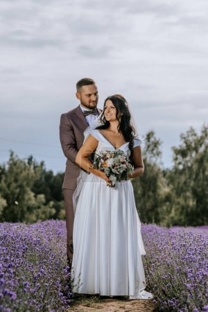 Photo for Valmiera, Latvia- July 28, 2024 - A couple in wedding attire stands amidst a field of lavender, with the groom gazing affectionately at the bride as she smiles and holds a bouquet. - Royalty Free Image