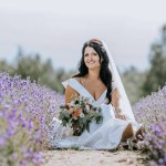 Valmiera, Latvia- July 28, 2023 - A radiant bride seated amidst a field of lavender looks up, her bouquet and smile bright under a sunny sky.