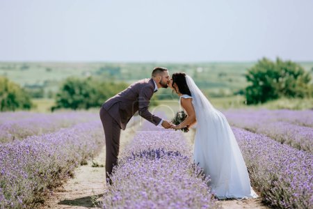 Photo for Valmiera, Latvia- July 28, 2024 -  a bride and groom are bending forward to share a kiss, holding hands in the middle of a lavender field with a scenic horizon in the background. - Royalty Free Image