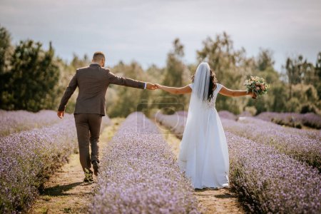 Foto de Valmiera, Letonia 28 de julio de 2024 - una novia y un novio tomados de la mano y caminando por un camino a través de un campo de lavanda, lo que sugiere un momento romántico y alegre después de la boda. - Imagen libre de derechos