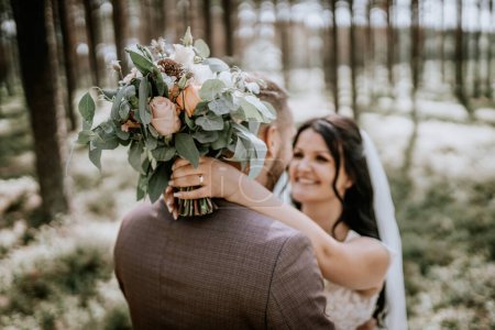Valmiera, Lettland - 28. Juli 2024 - Braut lächelt an einem Blumenstrauß vorbei, den der Bräutigam vor einem Waldhintergrund hält.
