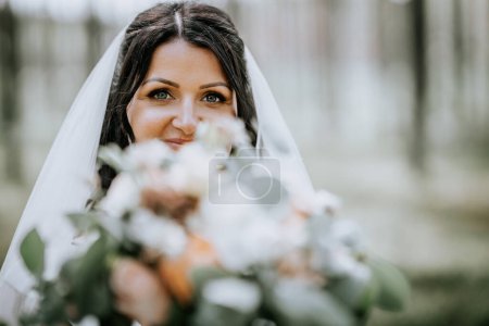 Photo for Valmiera, Latvia- July 28, 2024 - Bride peeks over her bouquet, veil draped, with a joyful gaze in a forest backdrop. - Royalty Free Image