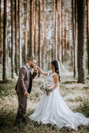 Valmiera, Letonia - 28 de julio de 2024 - Una novia y un novio se paran en un bosque, el novio besando la mano de la novia, ambos sonriendo calurosamente.