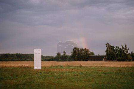 Valmiera, Latvia- July 28, 2024 - A standalone white door is set in an open field with a backdrop of a grey sky and distant trees, possibly suggesting a conceptual or surreal scene.