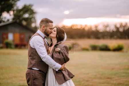 Valmiera, Latvia- July 28, 2024 - A loving couple dressed in wedding attire share a kiss in a field at dusk, with trees and a rustic building in the background.