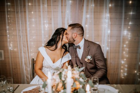 Valmiera, Latvia- July 28, 2024 - A bride and groom are kissing at a wedding table, with a white candle, a bouquet, and fairy lights in the background.