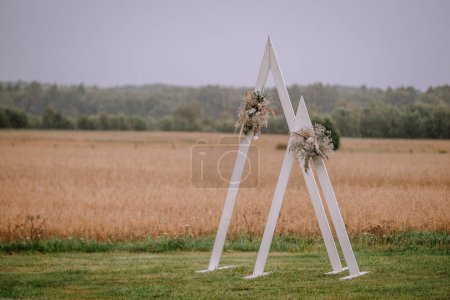 Photo for Valmiera, Latvia- July 28, 2024 - Large white letters 'A' and 'M' form an archway decorated with floral arrangements in a field under a rainy sky, creating a romantic, rustic wedding scene. - Royalty Free Image