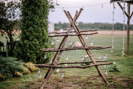 Valmiera, Latvia- July 28, 2024 - A rustic wooden shelf in an 'X' shape with hanging glass vases, set against an outdoor backdrop with greenery and a string of lights.