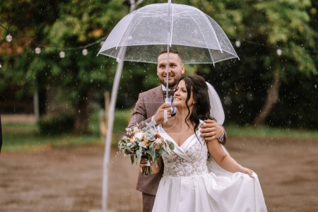 Photo for Valmiera, Latvia- July 28, 2024 - A joyful bride and groom share a moment under a transparent umbrella as rain gently falls around them. - Royalty Free Image