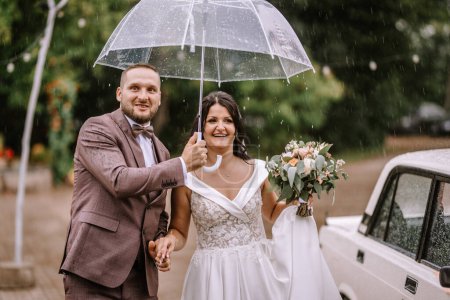 Photo for Valmiera, Latvia- July 28, 2024 - A newlywed couple smiles joyfully under a clear umbrella amidst a light shower, with a classic white car in the background. - Royalty Free Image