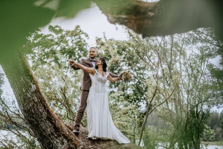 Photo for Valmiera, Latvia- July 28, 2024 - A bride and groom stand on a tree branch, extending their arms in a Titanic-like pose, surrounded by a leafy green canopy. - Royalty Free Image