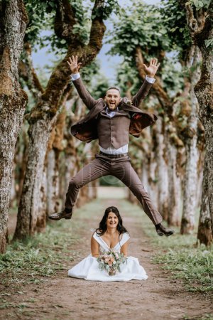 Valmiera, Lettland - 28. Juli 2024 - In einem skurrilen Hochzeitsmoment springt der Bräutigam hoch über die sitzende Braut, umrahmt von einer Allee majestätischer, moosbewachsener Bäume..