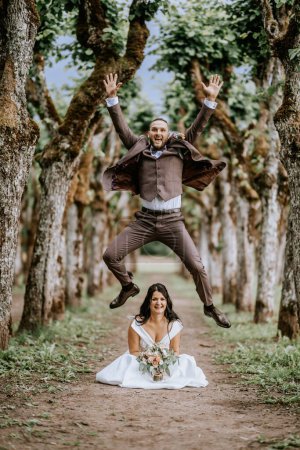 Valmiera, Lettland - 28. Juli 2023 - In einem skurrilen Hochzeitsmoment springt der Bräutigam hoch über die sitzende Braut, umrahmt von einer Allee majestätischer, moosbewachsener Bäume..