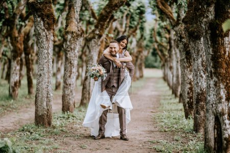 Valmiera, Lettland - 28. Juli 2023 - Ein Bräutigam, der seiner Braut in einem weißen Hochzeitskleid huckepack auf einem von alten, verdrehten Bäumen gesäumten Weg ein Lächeln schenkt, das Verspieltheit und Freude suggeriert.