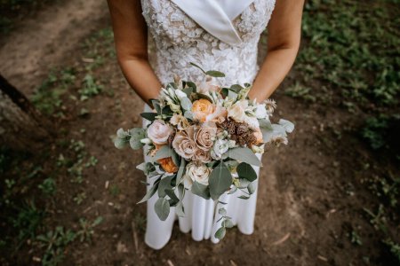 Photo for Valmiera, Latvia- July 28, 2023 - Close-up of a bride holding a bouquet with various flowers and greenery, in front of her lace-detailed wedding dress, set against an outdoor, earthy backdrop. - Royalty Free Image