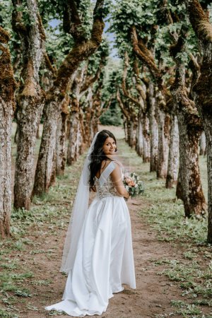 Valmiera, Lettonie- 28 juillet 2023 - Une mariée en robe blanche avec un corsage en dentelle et un long voile regarde par-dessus son épaule, tenant un bouquet, au milieu d'un bosquet d'arbres noueux