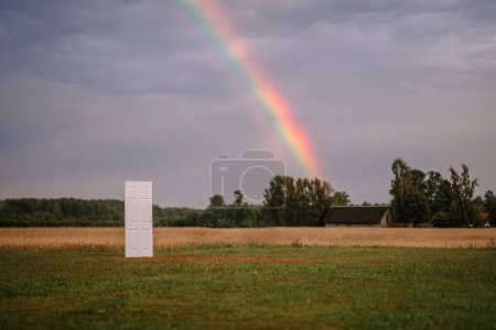 Valmiera, Latvia- July 28, 2024 - A standalone white door is set in an open field with a backdrop of a grey sky and distant trees, possibly suggesting a conceptual or surreal scene.