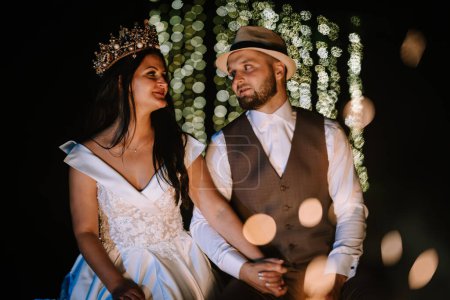 Valmiera, Latvia- July 28, 2023 - A bride wearing a tiara looks at the groom in a hat against a backdrop of bokeh lights.