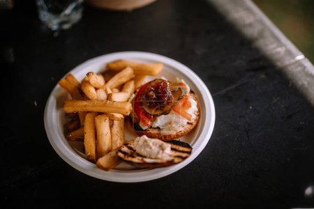 Valmiera, Latvia- July 29, 2023 - A burger with lettuce, tomato, and sauce alongside a portion of French fries served on a white disposable plate.