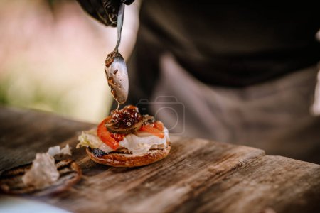 Valmiera, Latvia- July 29, 2023 - A burger being dressed with sauce from a spoon, tomatoes and lettuce on a bun, preparation in progress.