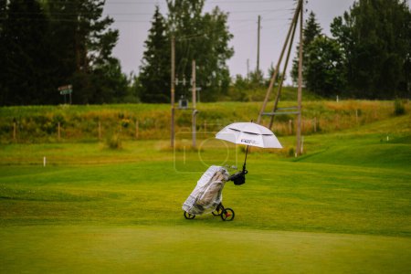 Foto de Sigulda, Letonia - 30 de julio de 2023 - Una bolsa de golf en un carro de tres ruedas con un paraguas blanco unido se encuentra en una exuberante calle verde bajo cielos nublados. - Imagen libre de derechos