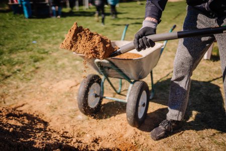 Valmiera, Latvia - April 21, 2024 - A person is shoveling soil into a wheelbarrow on a sunny day, showing a part of gardening or construction work.