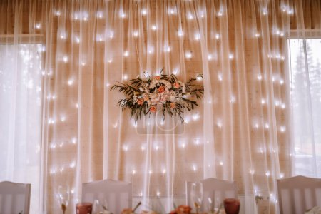 Valmiera, Lettonie - 5 août 2023 - Une toile de fond de mariage de rideaux transparents avec une chaîne de lumières de fées et un arrangement floral dans une salle lambrissée de bois.