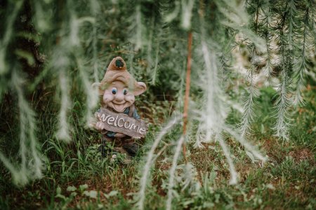 Valmiera, Latvia - August 5, 2023 - A garden gnome holding a "welcome" sign is nestled among green plants and grass, partially framed by hanging pine tree branches.