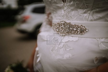 Téléchargez les photos : Valmiera, Lettonie - 5 août 2023 - Gros plan sur un détail de robe de mariée avec une ceinture de taille embellie, un tissu de dentelle et un fond flou avec une voiture blanche. - en image libre de droit
