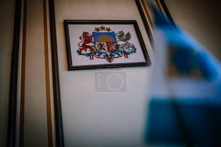 Foto de Valmiera, Letonia - 5 de agosto de 2023 - Un escudo de armas enmarcado en una pared, con un león, un grifo, un escudo con un rayo de sol y estrellas, y una cinta roja. Dresser de la República de Letonia - Imagen libre de derechos