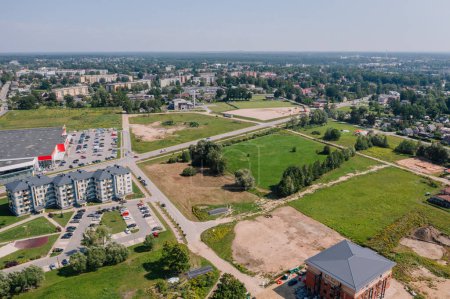 Valmiera, Latvia - August 7, 2023 - Aerial view of a suburban landscape with residential buildings, a large parking lot, green fields, and a main road.